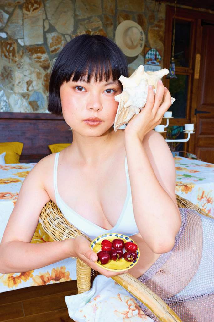 eine Frau in einem weißen Bikini mit einer Schale Kirschen in der Hand, in der anderen eine Muschel