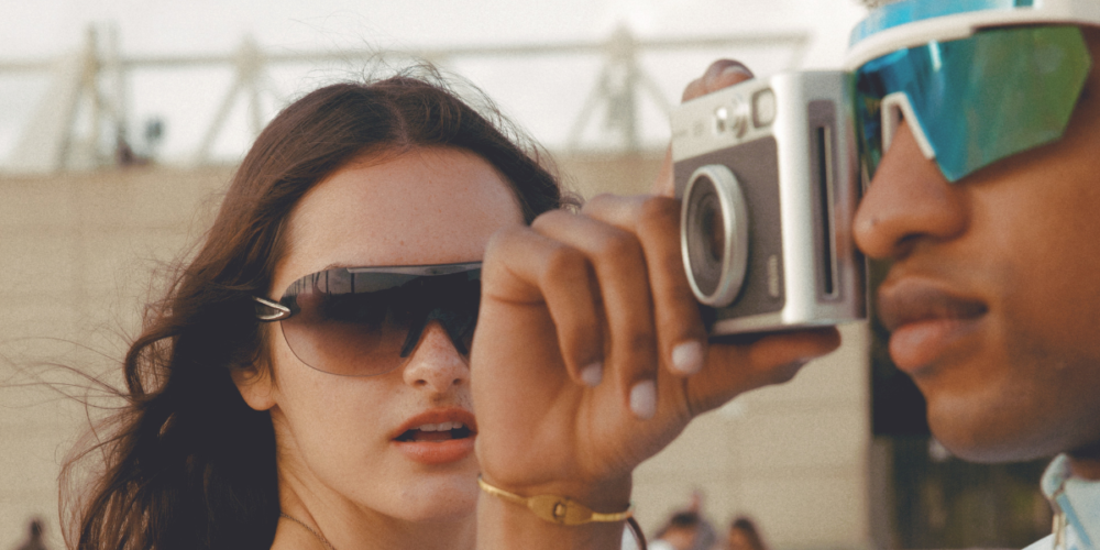 im Hintergrund eine junge Frau mit Sonnenbrille, im Vordergrund ein junger Mann der durch den Sucher einer Instax Kamera schaut